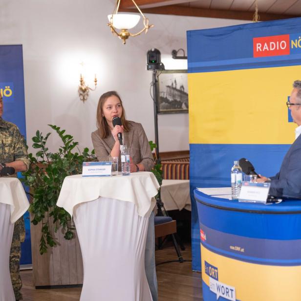 Drei Personen diskutieren an Stehtischen mit Mikrofonen und Namensschildern bei einer Radiosendung, Pflanzen im Hintergrund.