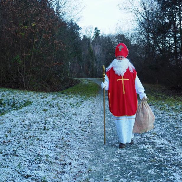 Eine als Nikolaus verkleidete Person mit rotem Mantel, Bischofsmütze und Stab geht auf einem frostigen Waldweg.