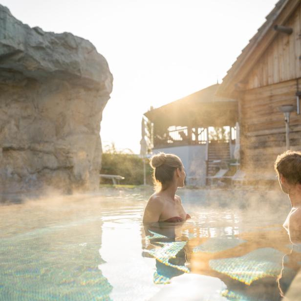Zwei Personen entspannen im dampfenden Außenpool vor einer Holzhütte bei Sonnenuntergang.