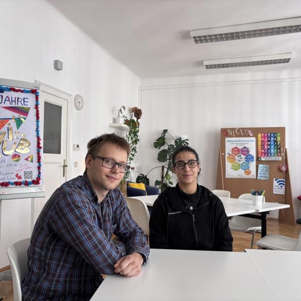 Zwei Personen sitzen an einem Tisch in einem hellen Raum mit bunten Plakaten und Pflanzen im Hintergrund.