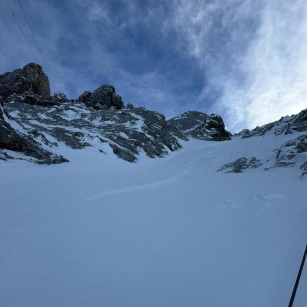Verschneite Felswand mit steilem Anstieg, im Vordergrund ein Sicherungsseil, darüber bewölkter Himmel.