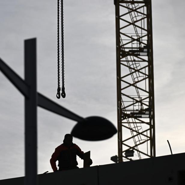 Eine Person arbeitet auf einer Baustelle neben einem Kran, im Vordergrund sind Straßenlaternen zu sehen.