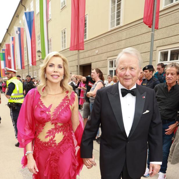 Ein elegant gekleidetes Paar geht Hand in Hand auf einer Veranstaltung im Freien, umgeben von Zuschauern und bunten Fahnen.