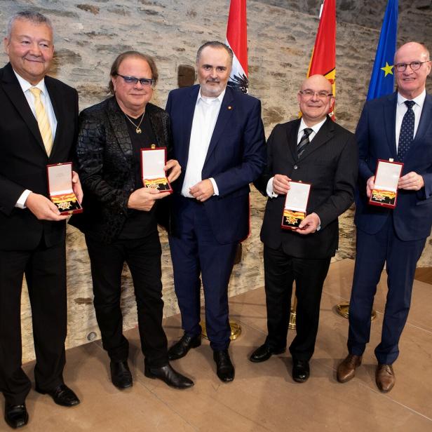 Fünf Männer in Anzügen stehen nebeneinander und halten Auszeichnungen vor sich, im Hintergrund sind Fahnen zu sehen.