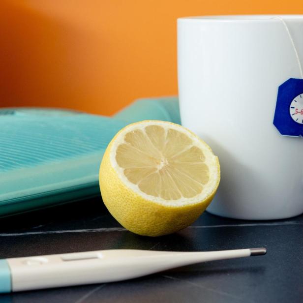 Tasse mit Teebeutel, halbe Zitrone, Wärmflasche und Fieberthermometer auf Tisch.