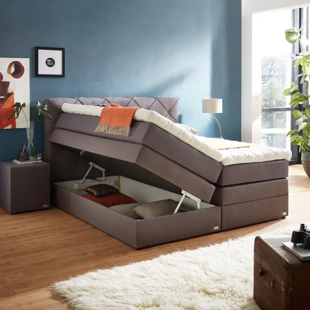 Hellgraues Polsterbett mit hochgeklapptem Lattenrost und Stauraum darunter, orangefarbene Decke, blaue Wand mit zwei Bildern, großer Fensterbereich mit Pflanzen.