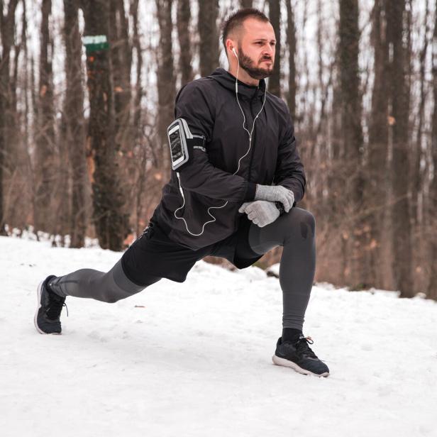 Ein Mann macht einen Ausfallschritt im verschneiten Wald und trägt Sportkleidung sowie Kopfhörer.