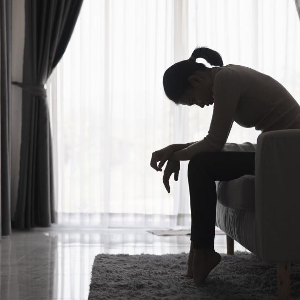 Eine Frau sitzt mit gesenktem Kopf und hängenden Schultern auf einem Sofa in einem abgedunkelten Raum.