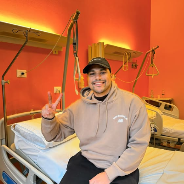 Infleuncer sitzt auf einem Krankenbett und hebt einen Daumen nach oben. 