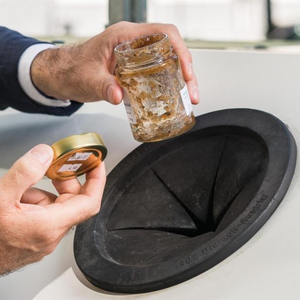 Eine männliche Hand hält ein leeres Marmeladeglas vor einem Glascontainer.