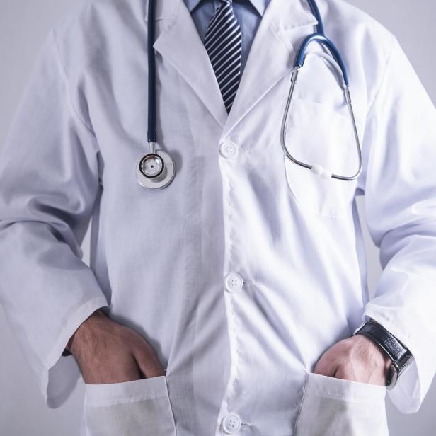 Ein Arzt im weißen Kittel mit Stethoskop und gestreifter Krawatte steht mit den Händen in den Taschen da.