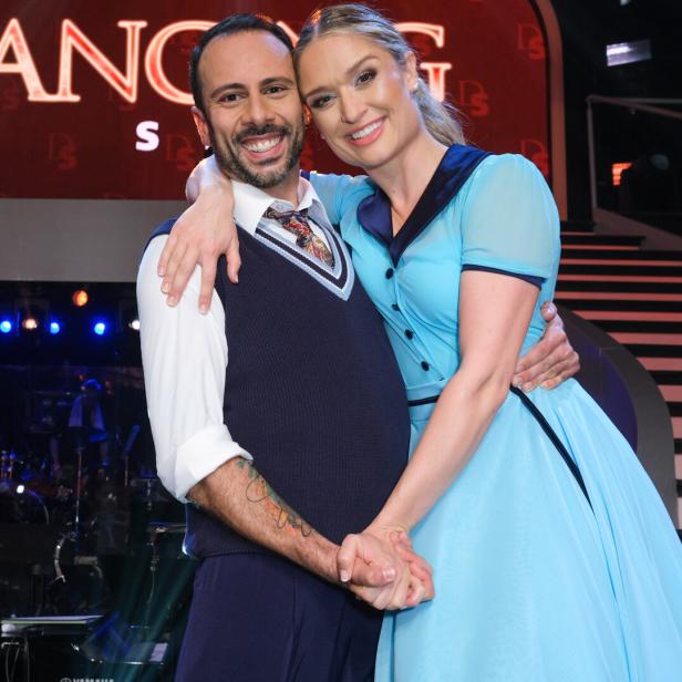 Ein Mann und eine Frau in eleganter Kleidung umarmen sich lächelnd auf einer Bühne mit Lichtern im Hintergrund.