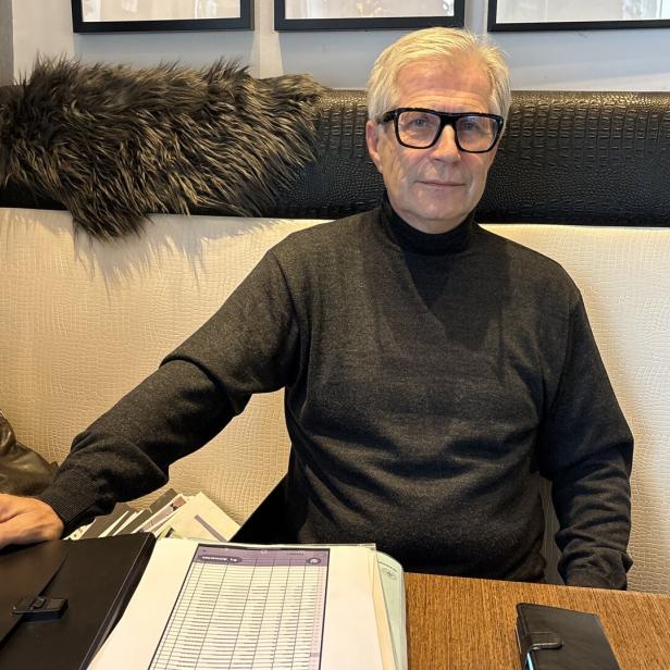 Ein älterer Mann mit Brille sitzt an einem Tisch mit Unterlagen und schaut freundlich in die Kamera.