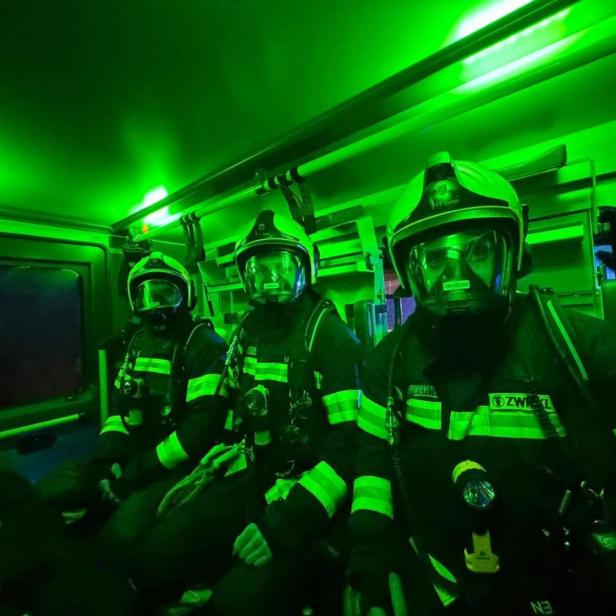Drei Feuerwehrleute in voller Schutzausrüstung sitzen in einem grün beleuchteten Einsatzfahrzeug.