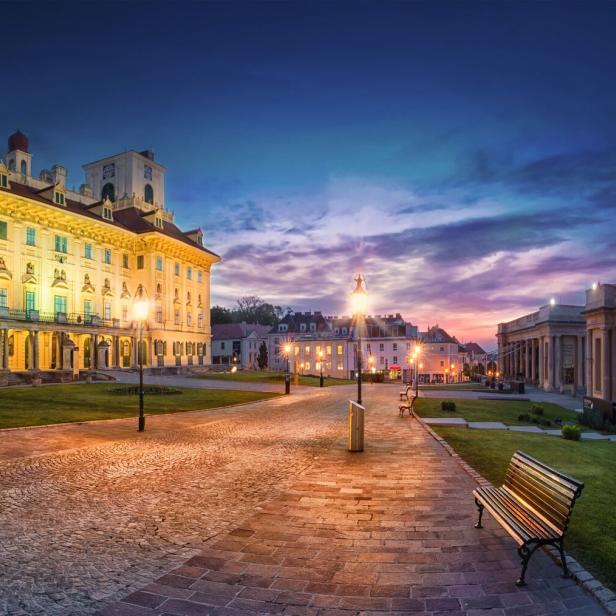 Historisches Schloss mit beleuchteter Fassade, Kopfsteinpflasterweg, Bänken und gepflegtem Garten bei Sonnenuntergang.