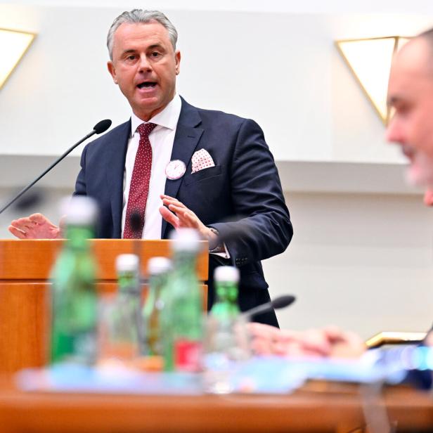 Ein Mann im Anzug spricht an einem Rednerpult, im Vordergrund sitzen weitere Personen mit Flaschen auf dem Tisch.