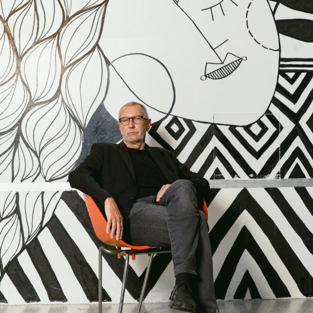 Konrad Paul Liessmann sitzt entspannt auf einem Stuhl vor einer schwarz-weiß gestalteten Wand mit abstrakten Mustern und Zeichnungen.