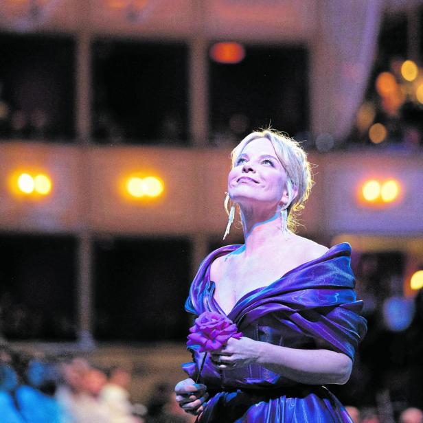 Eine elegant gekleidete Frau in einem lila Kleid hält eine Blume und blickt lächelnd nach oben in einem festlich beleuchteten Saal.