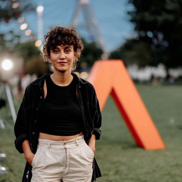 Junge Frau steht auf einer Wiese vor einer großen orangen Skulptur in A-Form.