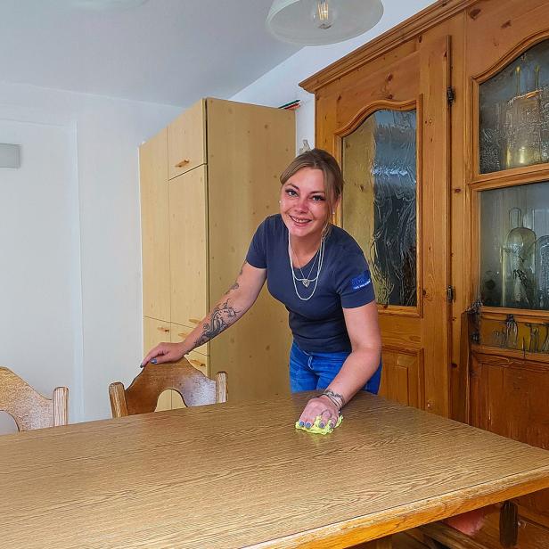 Eine Frau wischt lächelnd einen Holztisch in einem gemütlichen Esszimmer ab.