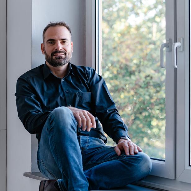 Mann mit Bart sitzt entspannt auf einer Fensterbank vor einem großen Fenster mit Blick ins Grüne.