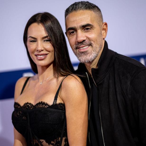 Eine Frau mit dunklem Haar und ein Mann mit Bart lächeln vor einem hellen Hintergrund.