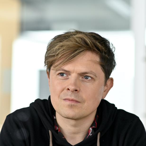 Ein Mann mit blond-braunen Haaren trägt einen schwarzen Hoodie und blickt nachdenklich zur Seite.