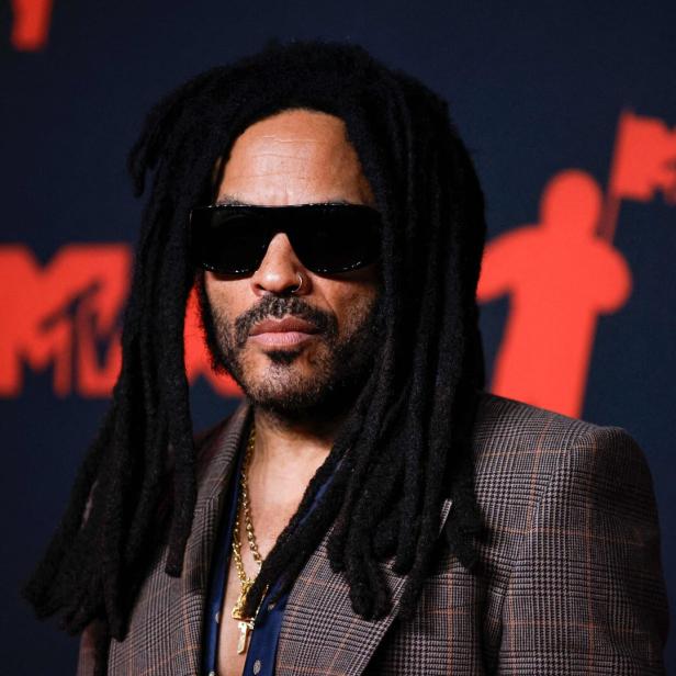 Eine Person mit langen Dreadlocks, Sonnenbrille und kariertem Sakko steht vor einem schwarzen Hintergrund mit roten Symbolen.