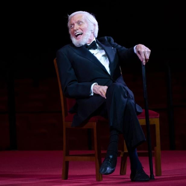 Ein älterer Mann im Smoking sitzt lachend auf einem Stuhl und hält einen Gehstock auf einer Bühne.