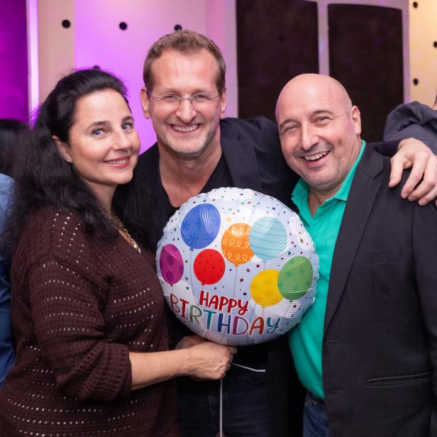 Drei lachende Menschen halten einen Geburtstagsballon mit bunten Luftballons und der Aufschrift „Happy Birthday“.