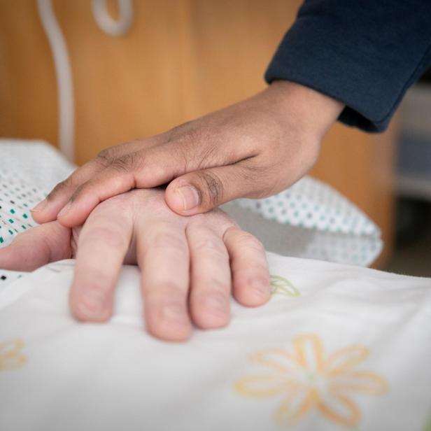 Eine Hand liegt tröstend auf einer anderen Hand, die auf einer Bettdecke mit Blumenmuster ruht.