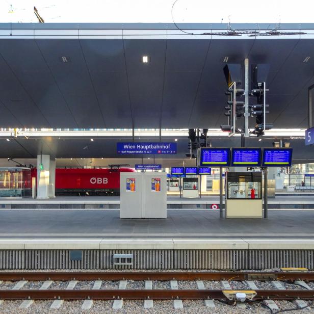 Bahnsteig am Wiener Hauptbahnhof mit Anzeigetafeln, rotem ÖBB-Zug und moderner Überdachung.