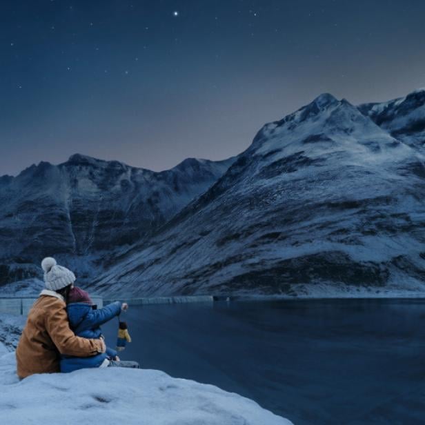Zwei Personen sitzen am verschneiten Ufer eines Stausees vor schneebedeckten Bergen bei Nacht.