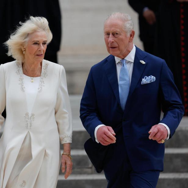 König Charles und Königin Camilla schreiten nebeneinander, er im blauen Anzug, sie in einem weißen Kleid.