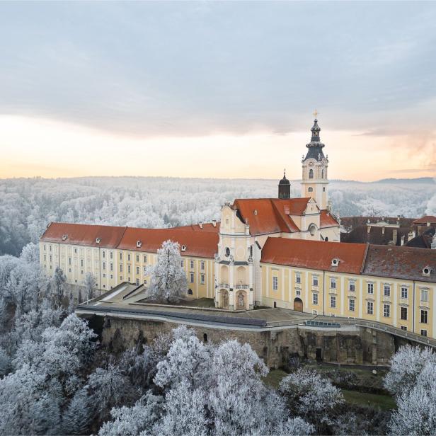 Das Stift Altenburg mit rotem Dach und Kirchturm liegt inmitten einer winterlichen, schneebedeckten Landschaft.