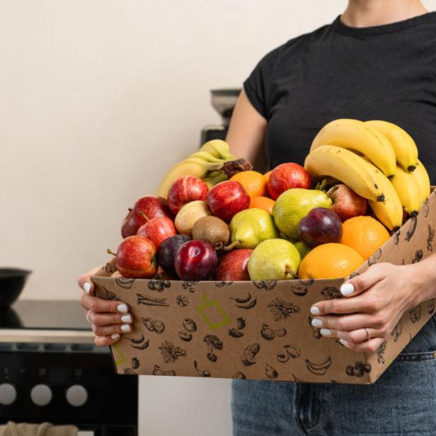 Eine frau in einem schwaren T-Shirt hält einen Obstkorb in den Händen und steht in einer Küche. 