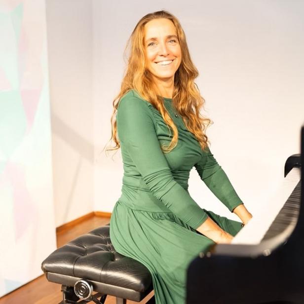 Frau in grünem Kleid sitzt lächelnd an einem schwarzen Flügel in hellem Raum.