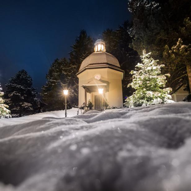 Verschneite kleine Kapelle bei Nacht, umgeben von beleuchteten Tannenbäumen und Laternen im Schnee.