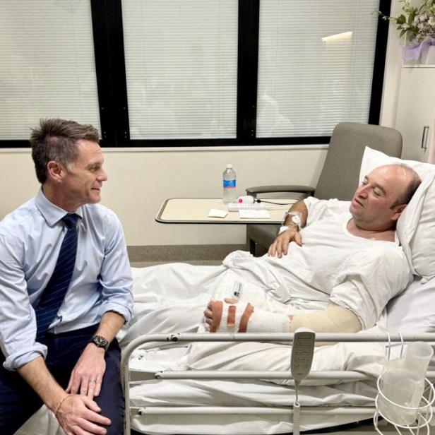 Ein Mann im Anzug sitzt am Krankenbett eines verletzten Mannes