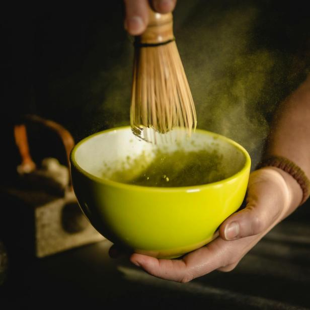 Hand hält eine gelbe Schale, in der mit einem Bambusbesen grünes Pulver aufgeschlagen wird.