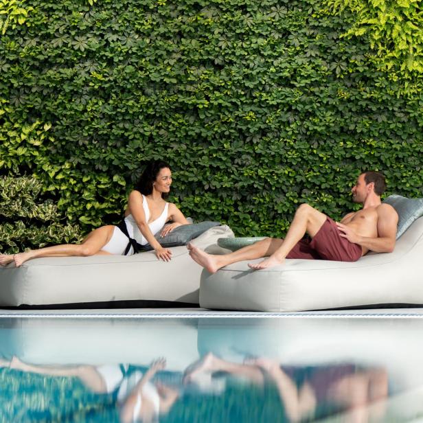 Zwei Personen entspannen auf Liegen am Pool vor einer grünen Pflanzenwand.