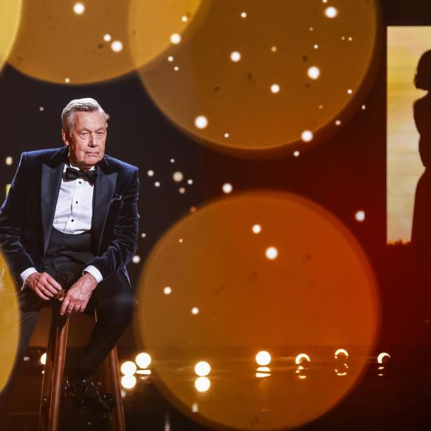 Ein älterer Mann im Smoking sitzt auf einem Hocker auf einer Bühne mit goldenen Lichtern und einer Silhouetten-Projektion im Hintergrund.