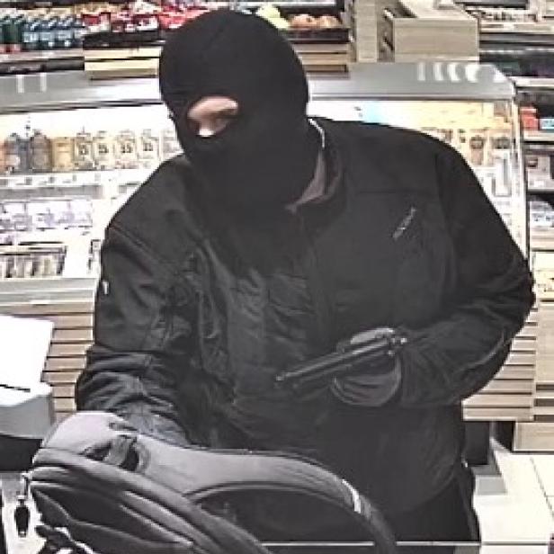 Ein maskierter Mann mit Sturmhaube, Bikerjacke hält eine Waffe in der Hand. Vor ihm steht ein Rucksack