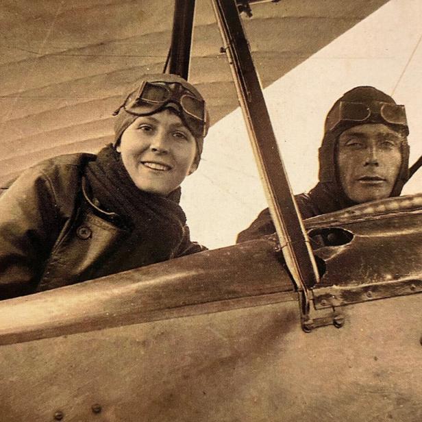 Zwei Personen mit Fliegermützen und Schutzbrillen sitzen in einem offenen Doppeldeckerflugzeug.