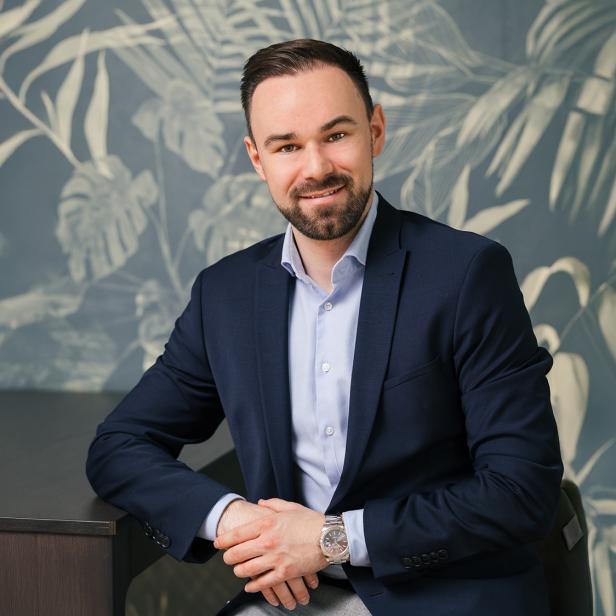 Mann im Anzug sitzt an einem Tisch vor einer Wand mit Pflanzenmuster und lächelt in die Kamera.