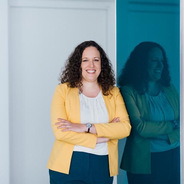 Frau mit lockigen Haaren und gelbem Blazer steht lächelnd vor einer Glaswand mit Spiegelung.