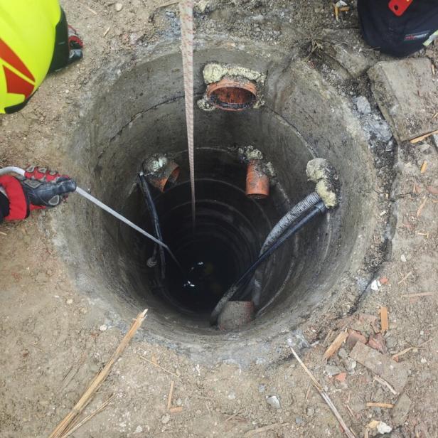 Zwei Personen in Schutzkleidung arbeiten an einem offenen, tiefen Schacht mit mehreren Rohren und Kabeln im Boden.