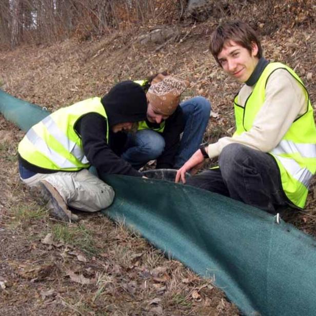 Drei Personen in Warnwesten befestigen am Straßenrand ein grünes Schutznetz am Boden.