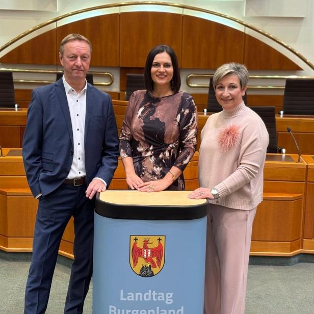Drei Personen stehen in einem Sitzungssaal hinter einem Rednerpult mit dem Wappen und Schriftzug „Landtag Burgenland“.