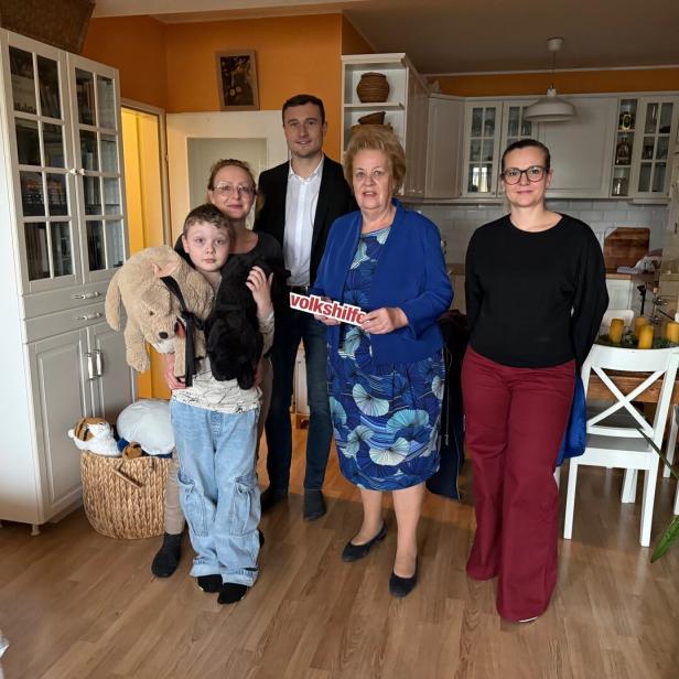 Fünf Personen stehen in einer Küche, ein Kind hält zwei Stofftiere, eine Frau hält ein Schild mit der Aufschrift „volkshilfe“.